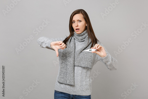 Puzzled young woman in gray sweater scarf showing thumb down, hold thermometer isolated on grey wall background. Healthy lifestyle, ill sick disease treatment, cold season concept. Mock up copy space. photo