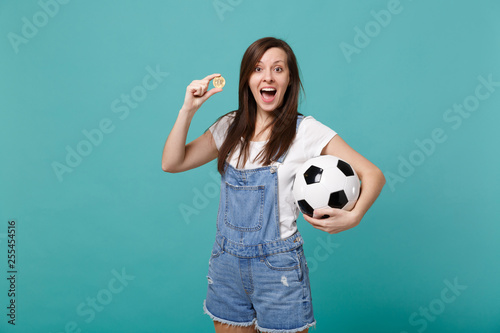 Surprised young woman football fan support favorite team with soccer ball, bitcoin future currency isolated on blue turquoise wall background. People emotions, sport family leisure lifestyle concept. © ViDi Studio