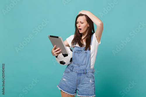 Puzzled young woman football fan holding soccer ball, using tablet pc computer putting hand on head isolated on blue turquoise wall background. People emotions, sport family leisure lifestyle concept.