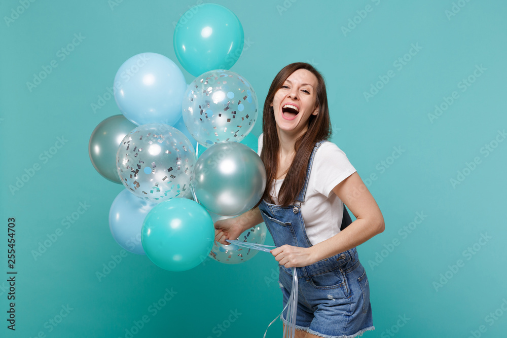 Fototapeta premium Portrait of cheerful laughing young woman in denim clothes celebrating and holding colorful air balloons isolated on blue turquoise wall background. Birthday holiday party, people emotions concept.