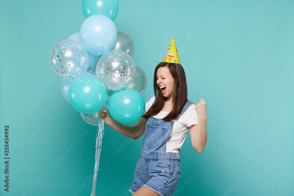 Fototapeta premium Overjoyed young woman in birthday hat screaming doing winner gesture, celebrating, holding colorful air balloons isolated on blue turquoise background. Birthday holiday party, people emotions concept.