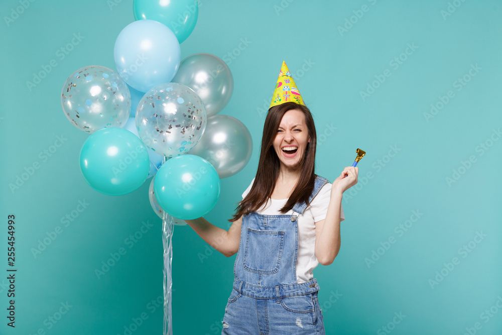 Fototapeta premium Laughing young woman in denim clothes, birthday hat holding pipe, celebrating with colorful air balloons isolated on blue turquoise wall background. Birthday holiday party, people emotions concept.