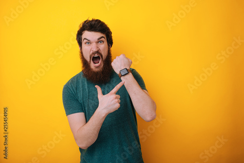 Young handsome bearded man In hurry pointing to watch time, impatience, upset and angry for deadline delay