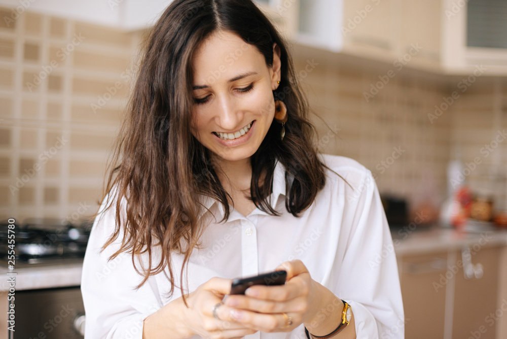 Obraz premium Beautiful young woman using her smartphone, texting and cheating standing in her kitchen