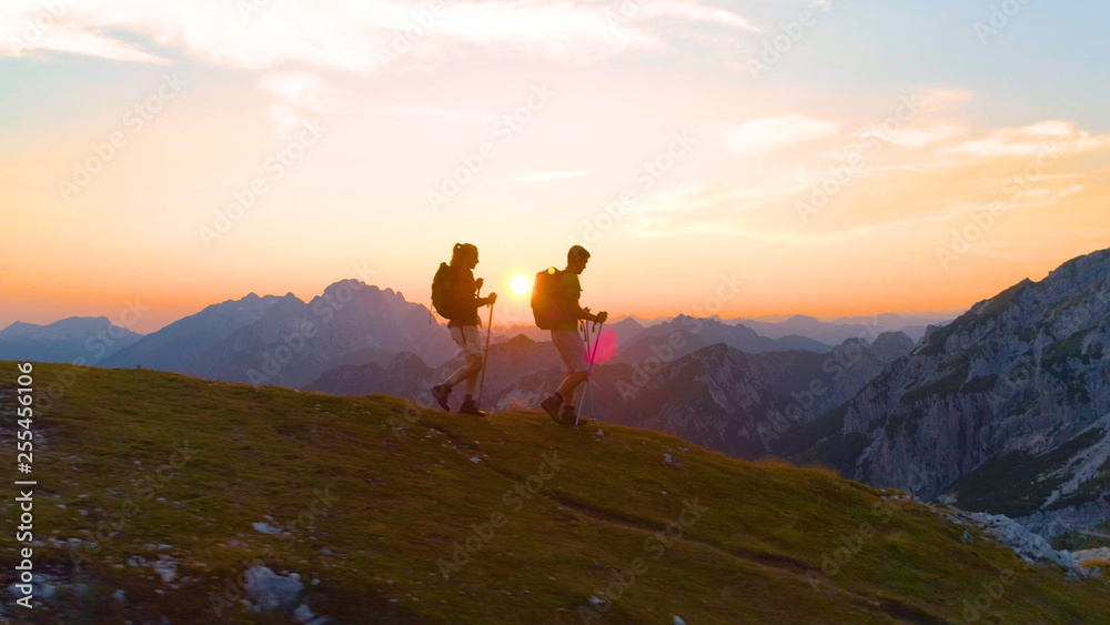 AERIAL: Flying along a cheerful couple hiking down a grassy hill in the Alps.