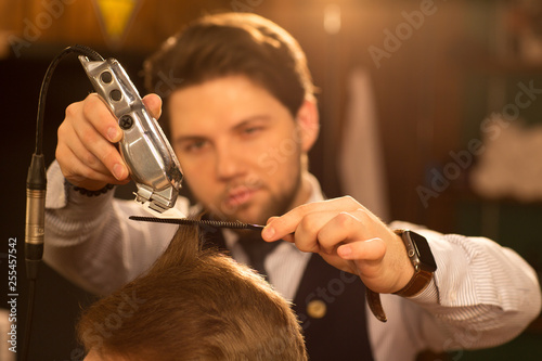Selective focus on a clipper trimmer in the hands of professional barber giving a haircut to his client working at the barbershop barbering service consumerism lifestyle fashion masculinity hipster ha