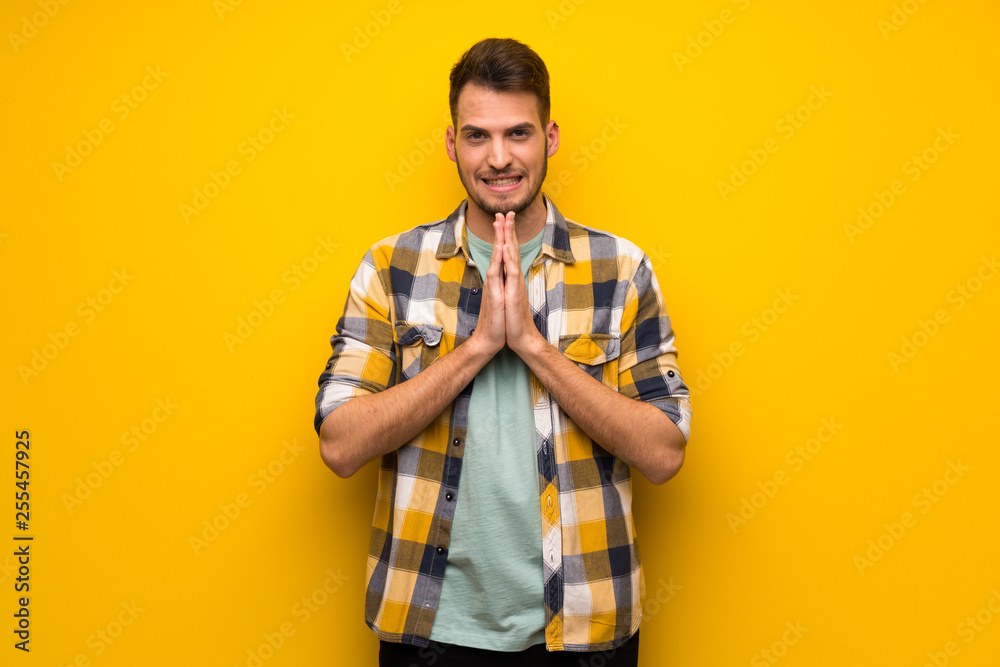 Handsome man over yellow wall keeps palm together. Person asks for something Handsome man over yellow wall keeps palm together. Person asks for something