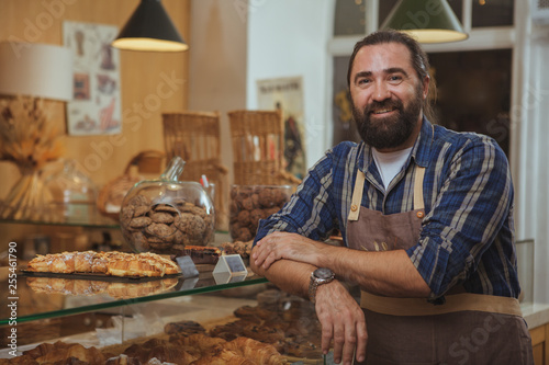 Tableau sur toile Handsome mature bearded male baker smiling to the camera proudly, working at his bakery shop, copy space