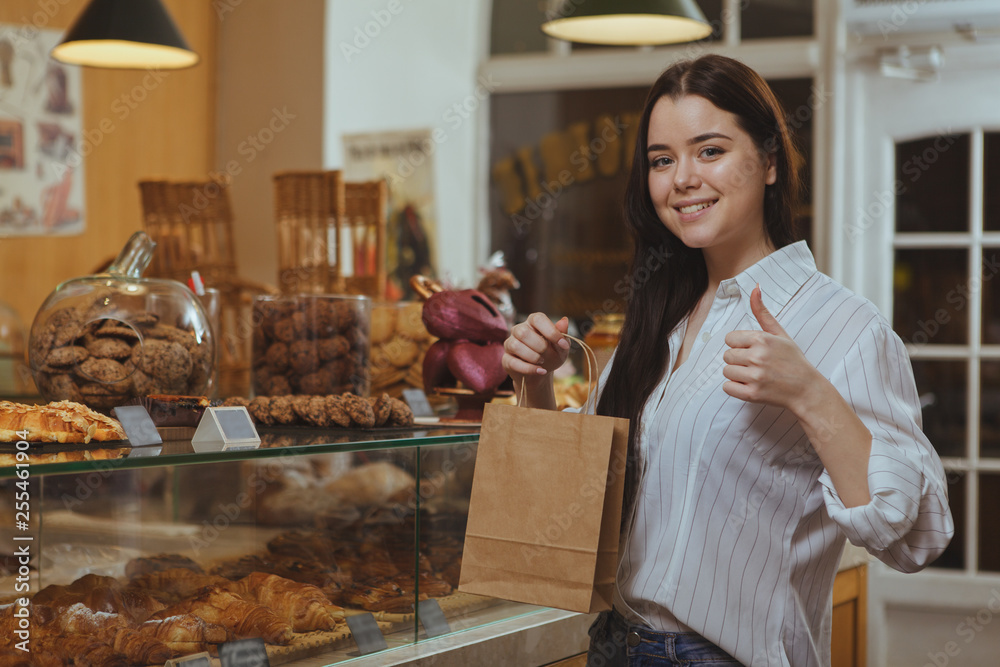Beautiful happy woman showing thumbs up, shopping at bakery with a bag in her hand. Attractive female customer buying food at local cafe, holding shopping bag. Retail, purchase concept