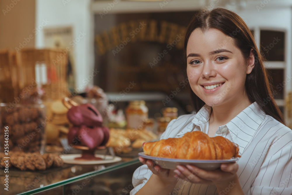 Close up of a beautiful happy young woman smiling cheerfully to the camera, holding delicious croissant on a plate, copy space. Lovely woman buying pastry at the bakery. Food, eating concept Close up of a beautiful happy young woman smiling cheerfully to the camera, holding delicious croissant on a plate, copy space. Lovely woman buying pastry at the bakery. Food, eating concept