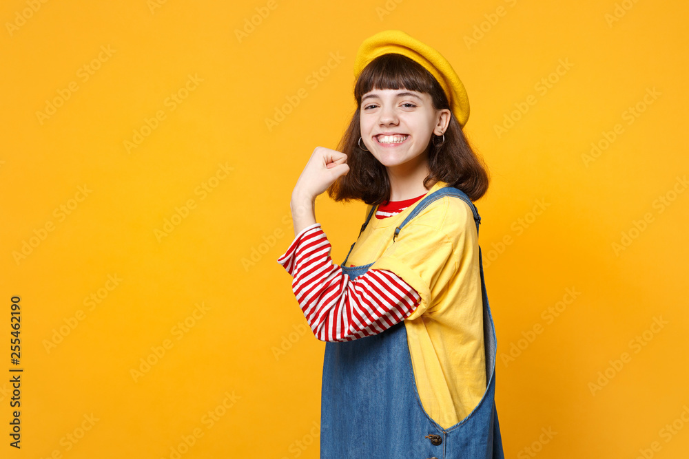 Portrait of smiling girl teenager in french beret, denim sundress showing biceps, muscles isolated on yellow wall background in studio. People sincere emotions, lifestyle concept. Mock up copy space. Portrait of smiling girl teenager in french beret, denim sundress showing biceps, muscles isolated on yellow wall background in studio. People sincere emotions, lifestyle concept. Mock up copy space.