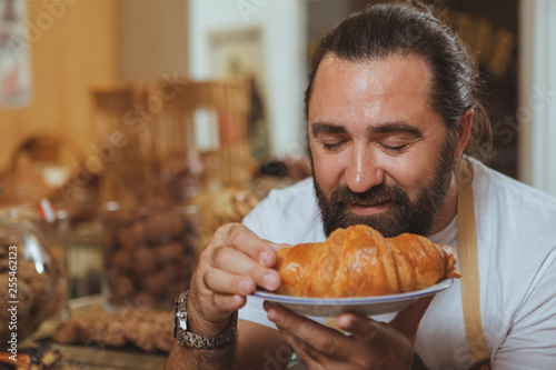 Close up portrait of a cheerful bearded male baker smelling delicious croissant  copy space. Handsome mature man working at the bakery  enjoying smell of freshly baked croissant. Selling pastry concep