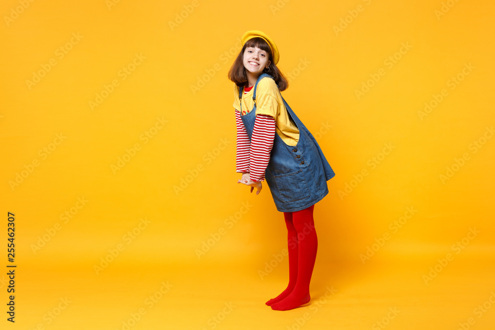 Full length portrait of beautiful girl teenager in french beret and denim sundress standing isolated on yellow wall background in studio. People sincere emotions lifestyle concept. Mock up copy space. Full length portrait of beautiful girl teenager in french beret and denim sundress standing isolated on yellow wall background in studio. People sincere emotions lifestyle concept. Mock up copy space.