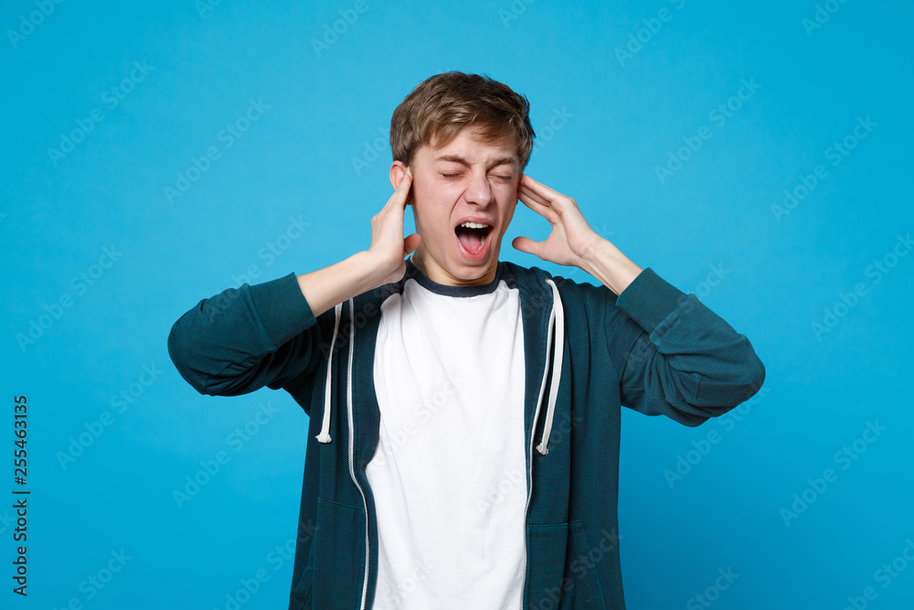 Portrait of screaming young man in casual clothes keeping eyes closed, covering ears with hands isolated on blue background in studio. People sincere emotions, lifestyle concept. Mock up copy space. Portrait of screaming young man in casual clothes keeping eyes closed, covering ears with hands isolated on blue background in studio. People sincere emotions, lifestyle concept. Mock up copy space.