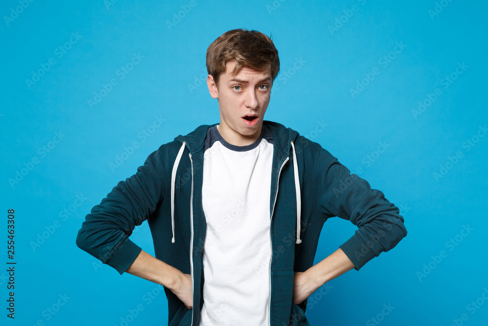 Portrait of strict young man in casual clothes standing with arms akimbo on waist, swearing isolated on blue wall background in studio. People sincere emotions, lifestyle concept. Mock up copy space. Portrait of strict young man in casual clothes standing with arms akimbo on waist, swearing isolated on blue wall background in studio. People sincere emotions, lifestyle concept. Mock up copy space.