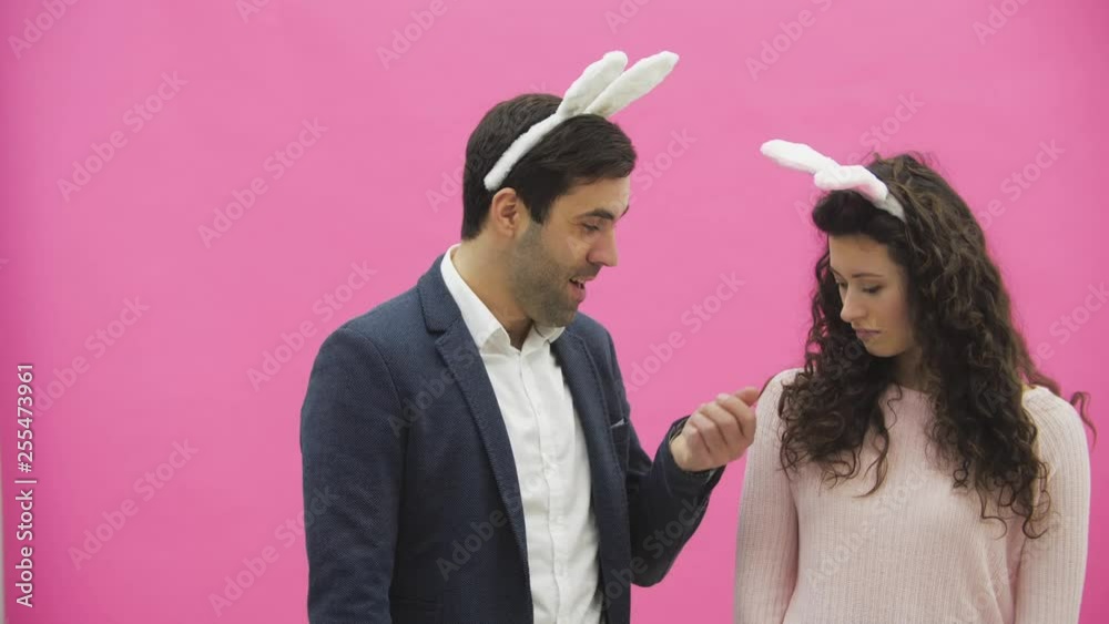 Young couple are beautiful on pink background. During this time, they are dressed in rabble ears. Looking at each other. A man shows carrots, his wife does not like the size of carrots and she throws Young couple are beautiful on pink background. During this time, they are dressed in rabble ears. Looking at each other. A man shows carrots, his wife does not like the size of carrots and she throws