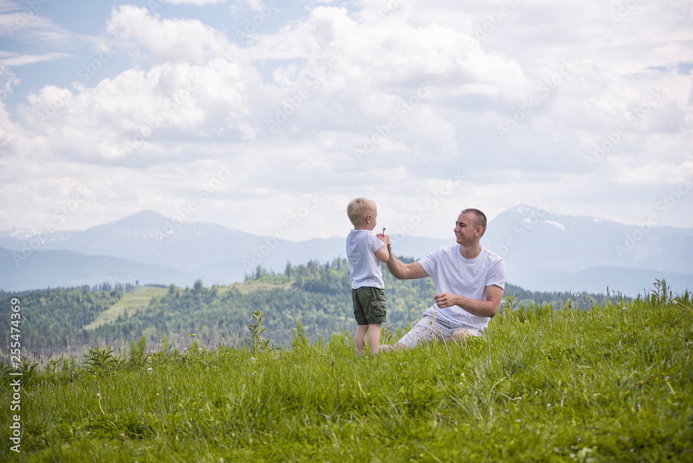 Father and young son are blowing dandelions sitting in the grass on a background of green forest, mountains and sky with clouds. Friendship concept Father and young son are blowing dandelions sitting in the grass on a background of green forest, mountains and sky with clouds. Friendship concept