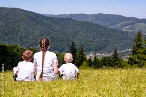 Mother and two young sons sitting on grass on the background of coniferous forest and mountains