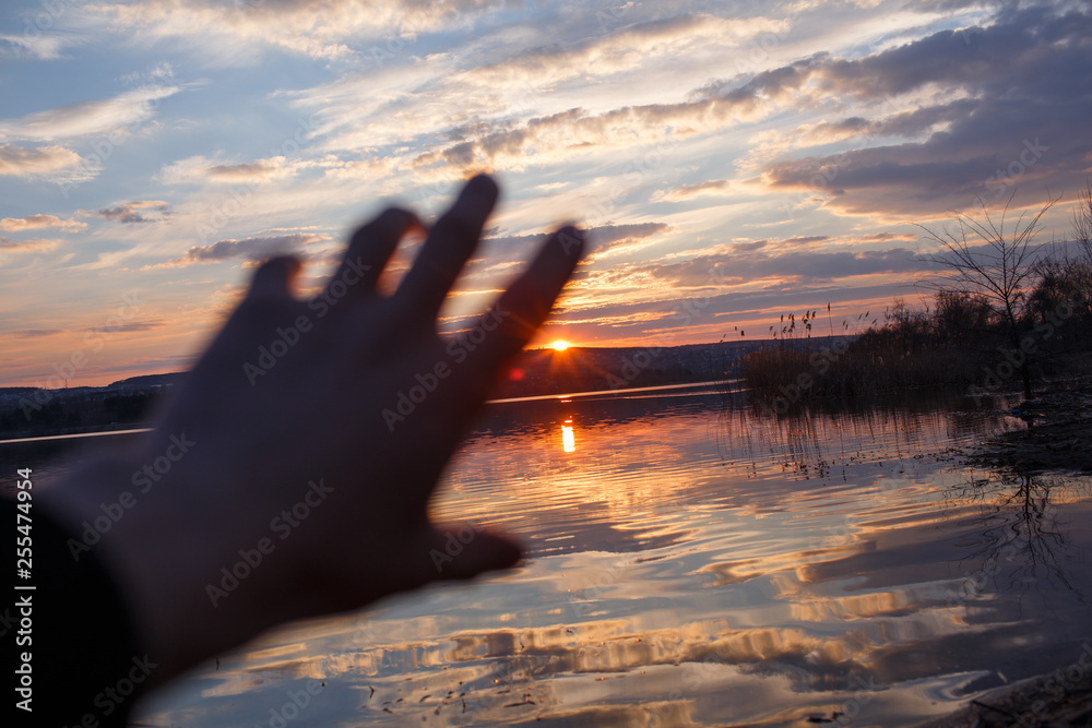 Male hand is touching the sunset over lake.