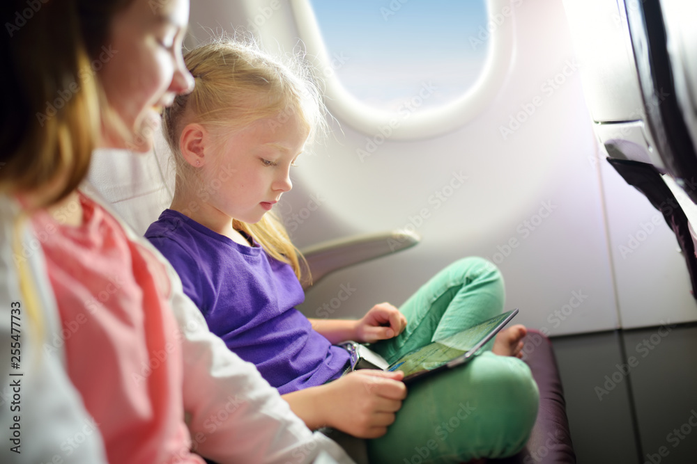 Adorable little girls traveling by an airplane. Children sitting by aircraft window and using a digital tablet during the flight. Traveling with kids.