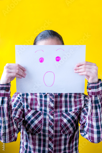 Girl in plaid shirt on yellow background holding white sheet of paper with a drawn smile of surprise