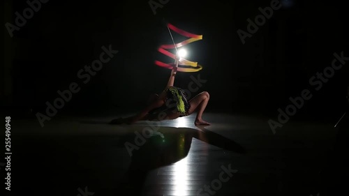 Slow motion, slim cute attractive girl athlete in bright colorful swimsuit performs elements of rhythmic gymnastics with ribbon in dark gym with beautiful staged light and contour light