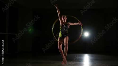 Slow motion, slim cute attractive girl athlete in bright colorful swimsuit performs elements of rhythmic gymnastics with hoop in dark gym with beautiful staged light and rear light