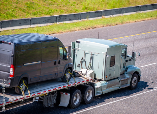 Powerful big rig classic semi truck transporting mini van fastened on flat bed semi trailer running on wide highway