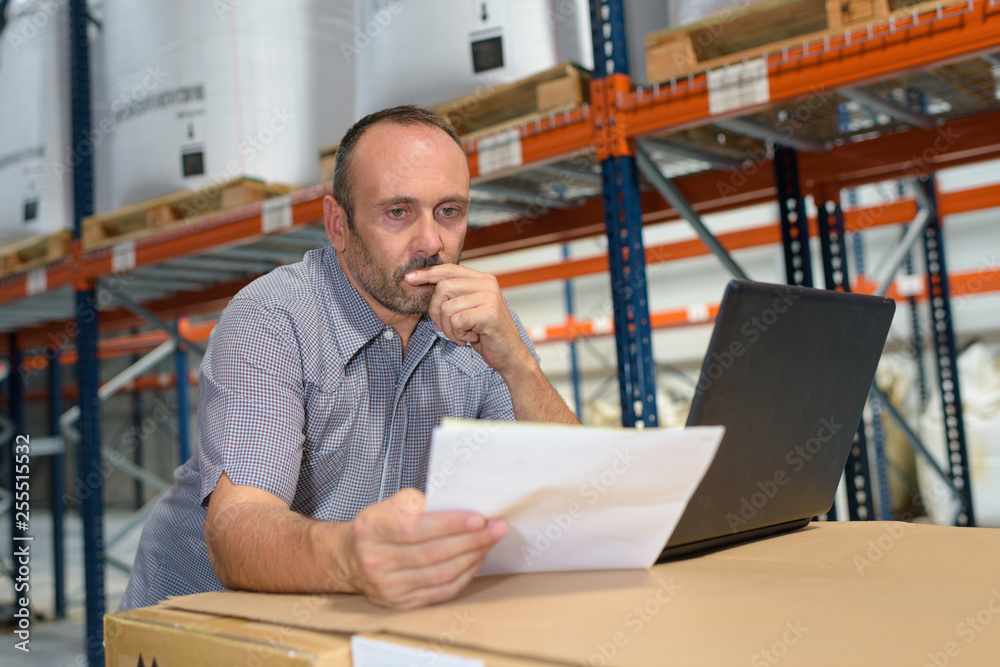 man in warehouse with laptop looking at papers