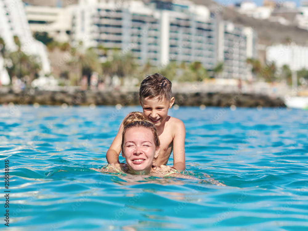 Cute European boy is sitting on his mother’s back, while swimming in the sea.