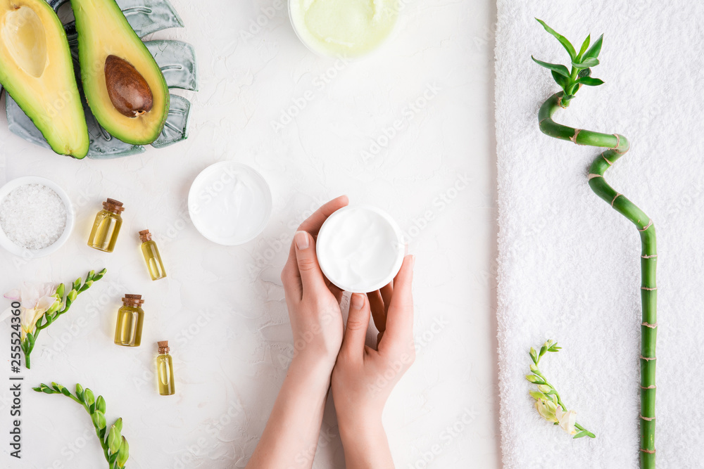 Top view and flat lay of woman holding cream on hands over white table with cosmetic products - avocado oil, cream and bamboo Top view and flat lay of woman holding cream on hands over white table with cosmetic products - avocado oil, cream and bamboo