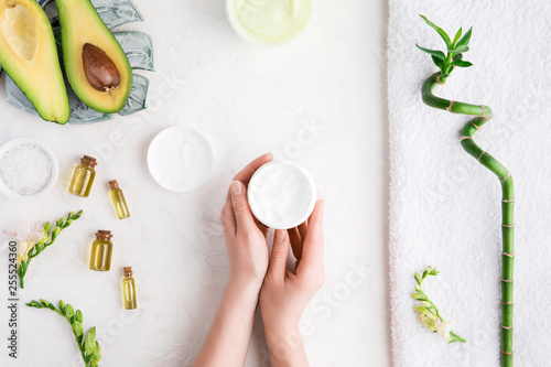 Top view and flat lay of woman holding cream on hands over white table with cosmetic products - avocado oil, cream and bamboo