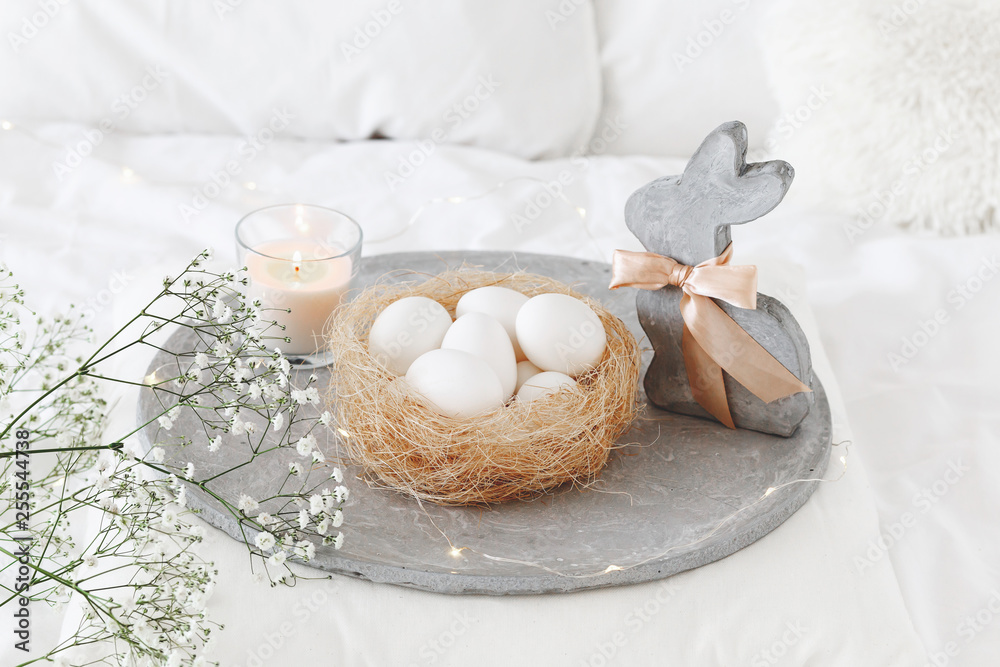 White modern bedroom with Easter decoration. Bed with white bedding set, pillows, concrete tray, nest with white eggs, decorative bunny figure, candle and gypsophila flowers. White modern bedroom with Easter decoration. Bed with white bedding set, pillows, concrete tray, nest with white eggs, decorative bunny figure, candle and gypsophila flowers.