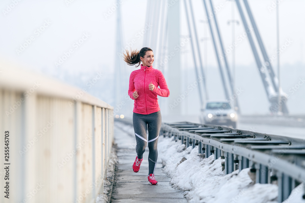 Pretty Caucasian woman with ponytail dressed in sportswear running on the bridge at winter. Healthy lifestyle concept.