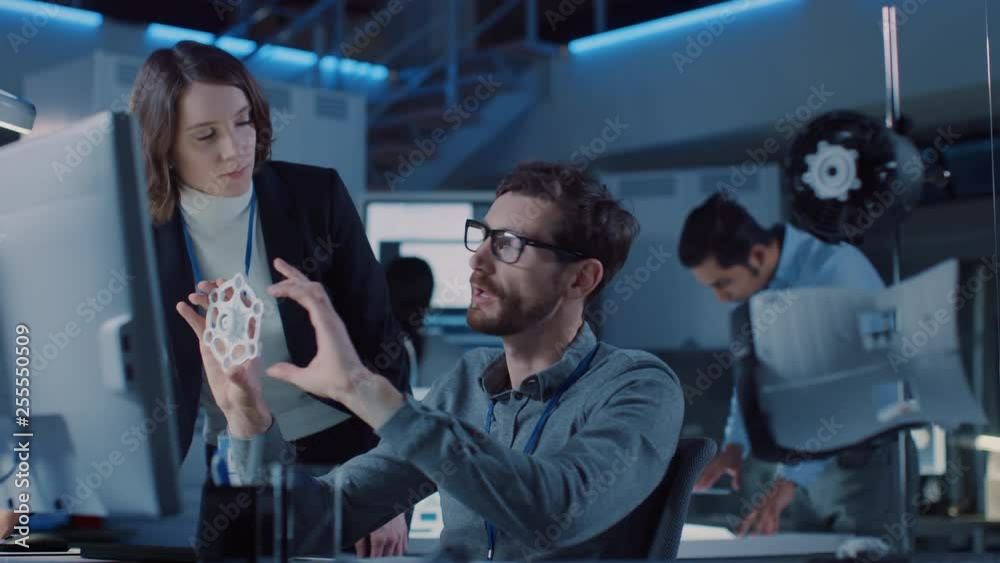 Futuristic Machine Engine Development Engineer Working on Computer at His Desk, Talks with Female Project Manager. Team of Professionals Working in the Modern Industrial Design Facility Futuristic Machine Engine Development Engineer Working on Computer at His Desk, Talks with Female Project Manager. Team of Professionals Working in the Modern Industrial Design Facility