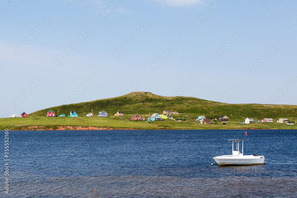 Obraz premium Small white boat anchored in water with scattering of colorful houses on Magdalen Islands shore, Quebec, Canada 