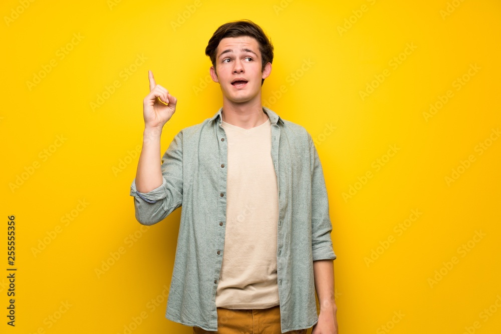 Teenager man over yellow wall intending to realizes the solution while lifting a finger up Teenager man over yellow wall intending to realizes the solution while lifting a finger up