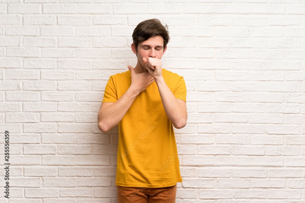 Teenager man over white brick wall is suffering with cough and feeling bad Teenager man over white brick wall is suffering with cough and feeling bad
