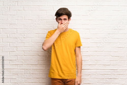 Teenager man over white brick wall covering mouth with hands for saying something inappropriate