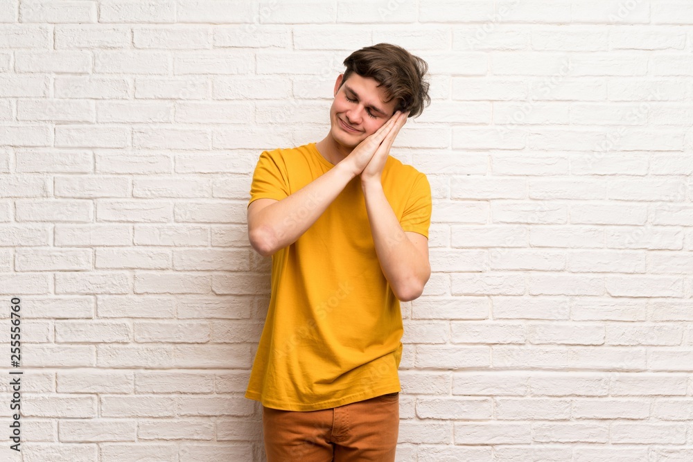Teenager man over white brick wall making sleep gesture in dorable expression Teenager man over white brick wall making sleep gesture in dorable expression
