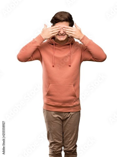 Teenager man with sweatshirt covering eyes by hands. Surprised to see what is ahead over isolated white background