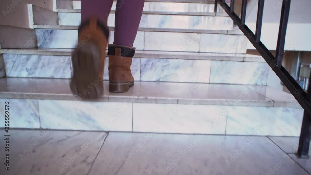 A young woman in beige leggings and brown boots climbs the marble steps to the indoors. The stone staircase is decorated with metal black railing. A young woman in beige leggings and brown boots climbs the marble steps to the indoors. The stone staircase is decorated with metal black railing.