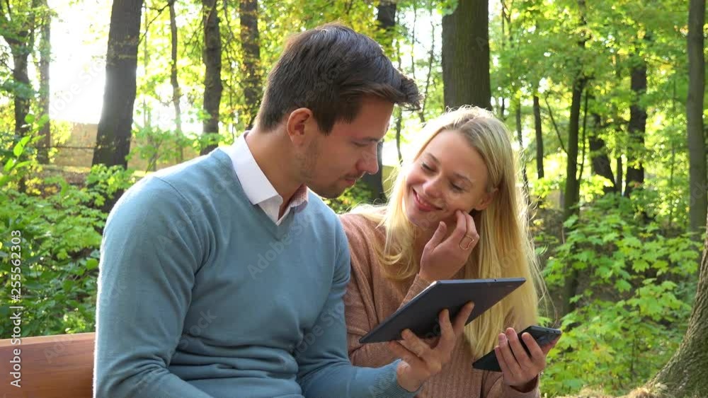 A young attractive couple works on a smartphone and a tablet on a bench in a park on a sunny day - closeup A young attractive couple works on a smartphone and a tablet on a bench in a park on a sunny day - closeup