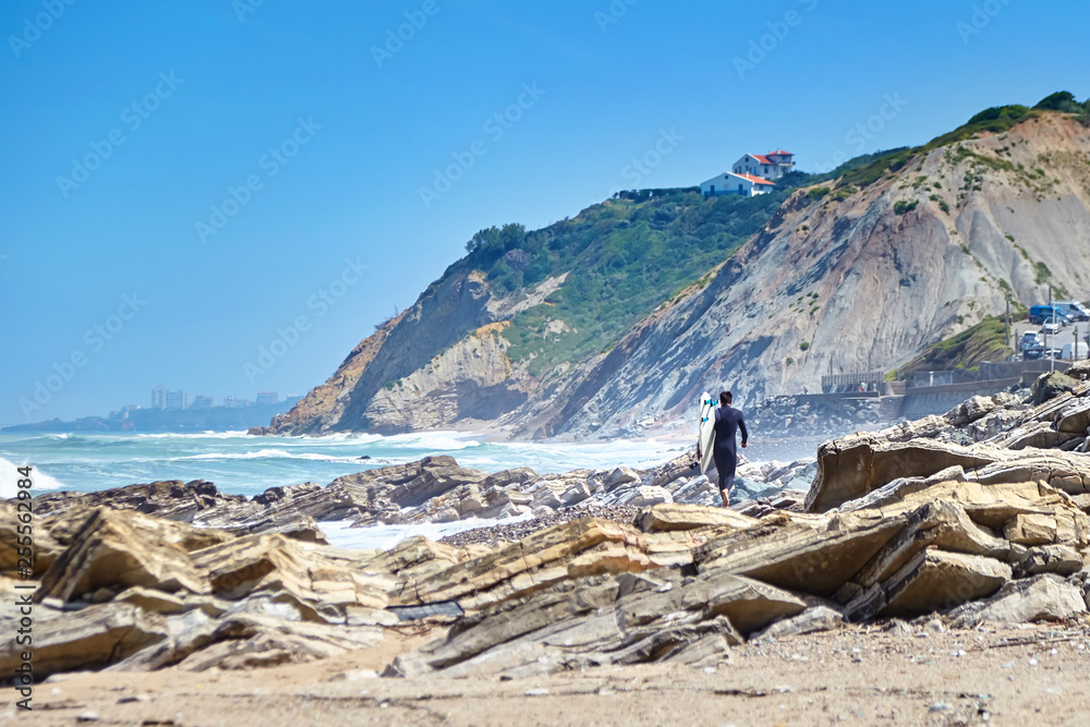 Surfer walks on rocky beach with surfboard. Atlantic ocean coast. Bidart, France