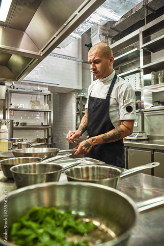 Restaurant kitchen. Vertical photo of concentrated chef in apron with several tattoos on his arms holding a frying pan