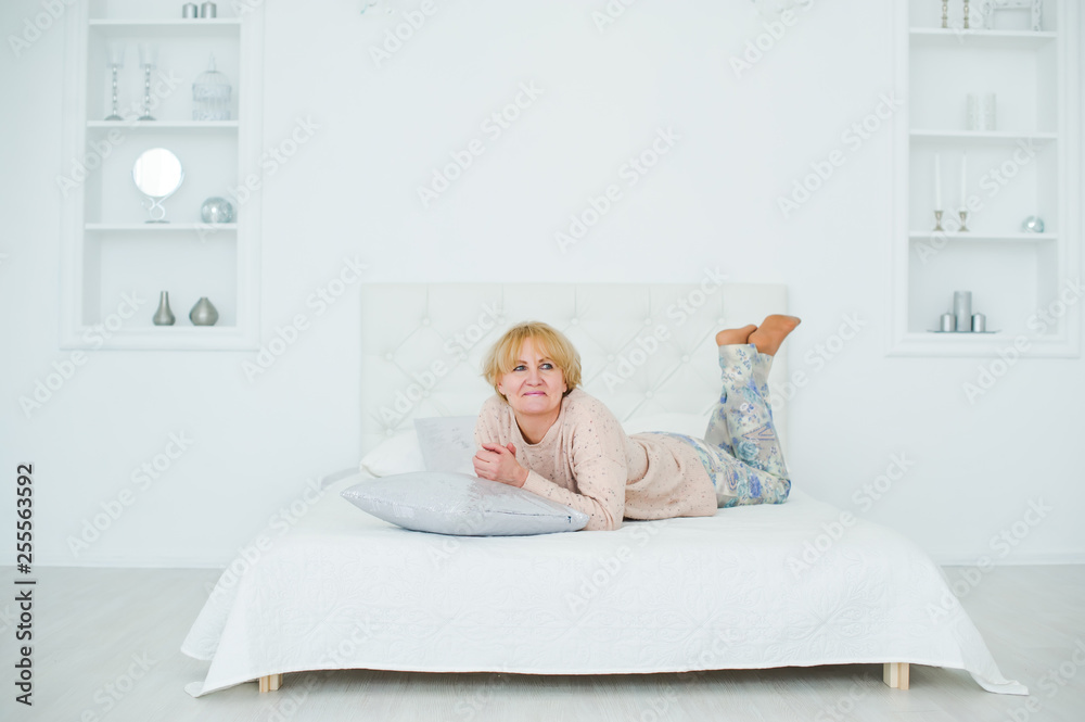 Smiling middle-aged attractive woman lying on white bed at home Smiling middle-aged attractive woman lying on white bed at home