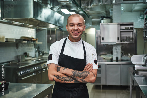 I love my work Cheerful young chef in apron keeping tattooed arms crossed and smiling while standing in a restaurant kitchen photo