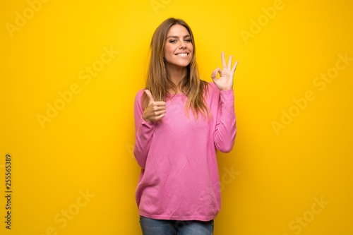 Woman with pink sweater over yellow wall showing ok sign with and giving a thumb up gesture