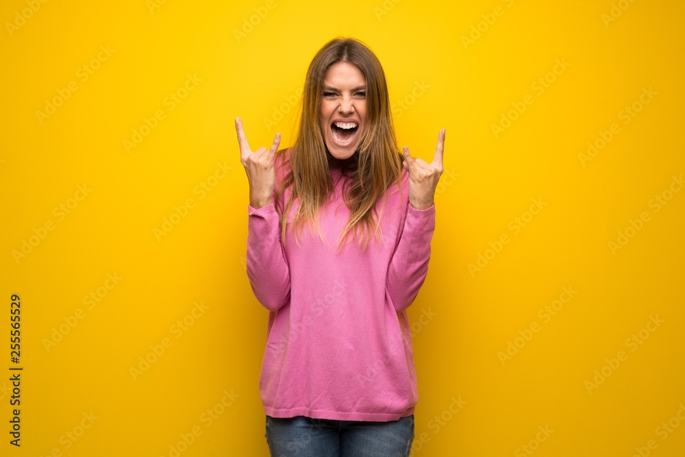 Woman with pink sweater over yellow wall making rock gesture Woman with pink sweater over yellow wall making rock gesture
