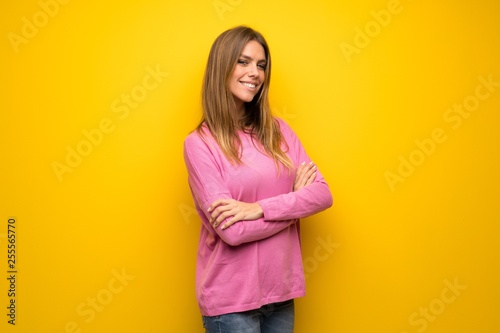 Woman with pink sweater over yellow wall with arms crossed and looking forward